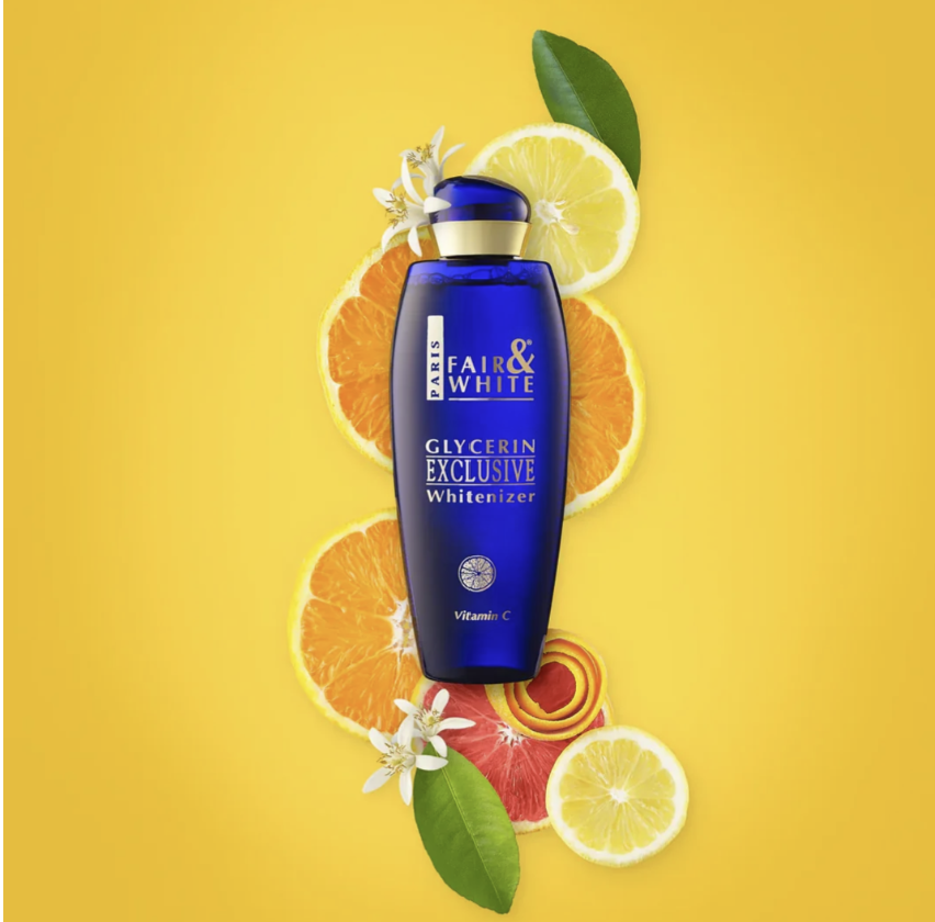 A bottle of Fair & White Glycerin Exclusive Whitizer in a deep blue bottle with a gold cap, styled on a bright yellow background with citrus slices, grapefruit slices, and green leaves.