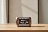 Thumbnail for Bar of African Black Soap wrapped in clear plastic with 'Reviv2Grow' branding on a wooden surface.