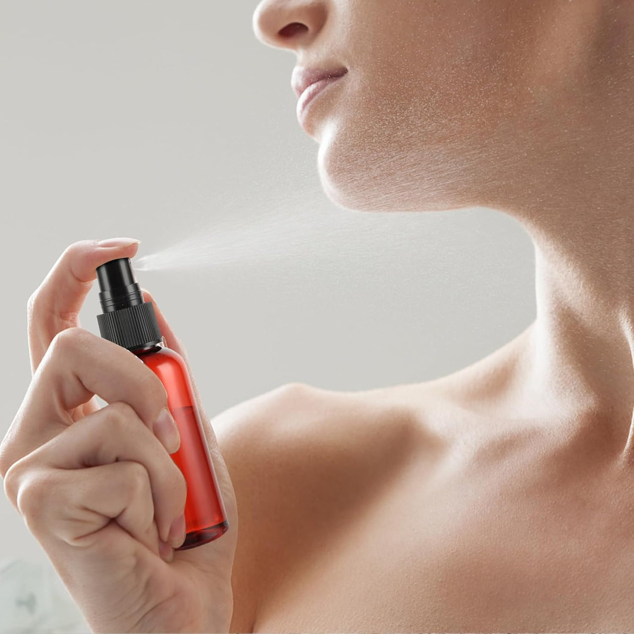 A close-up of a woman spraying a red and black perfume or body spray bottle on her neck and shoulder, with the mist visible against a soft light gray background.