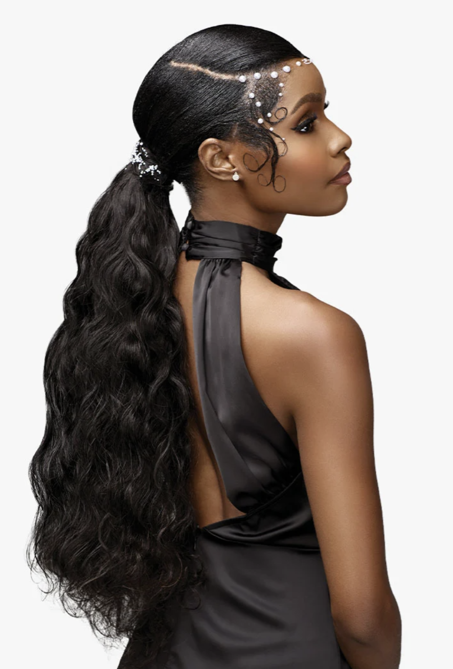 A woman shown from the back and side wearing a long, dark wavy ponytail extension attached to her hair, styled with a jeweled headpiece and pearl earrings against a light gray studio background.